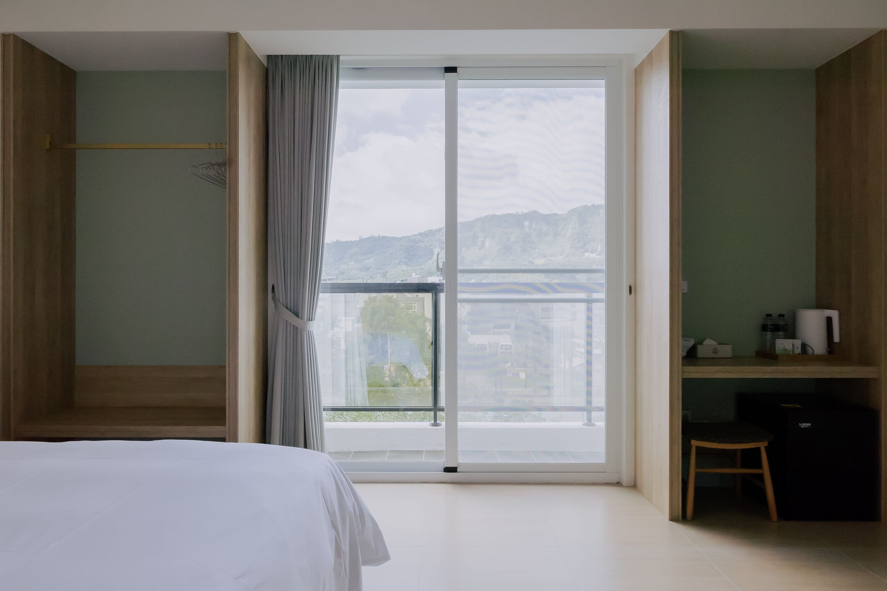 A bedroom with a large sliding glass door leading to a balcony, a bed, a desk with a chair, and a small cabinet.