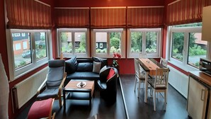 A modern living room with a black leather sofa, a wooden coffee table, and a dining table with chairs. Large windows with striped curtains provide views of the outside. - Charmantes Refugium, Harz (Wildemann)