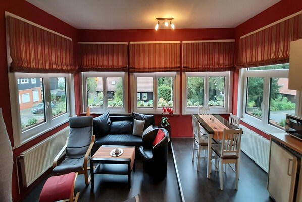 A modern living room with a black leather sofa, a wooden coffee table, and a dining table with chairs. Large windows with striped curtains provide views of the outside.