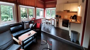 A modern living room with a sofa, dining table, and chairs. There are large windows with curtains, a kitchen area with appliances, and a trash bin. - Charmantes Refugium, Harz (Wildemann)