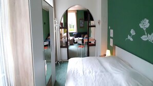 Ein Hotelzimmer mit Bett, Spiegel, grüner Wand mit Rosenmuster und Blick auf den Speisesaal durch einen Bogen.