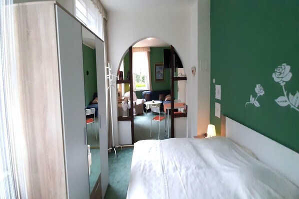 Quarto de hotel com cama, espelho, parede verde com padrão de rosa e vista para a sala de jantar através de uma porta arqueada.