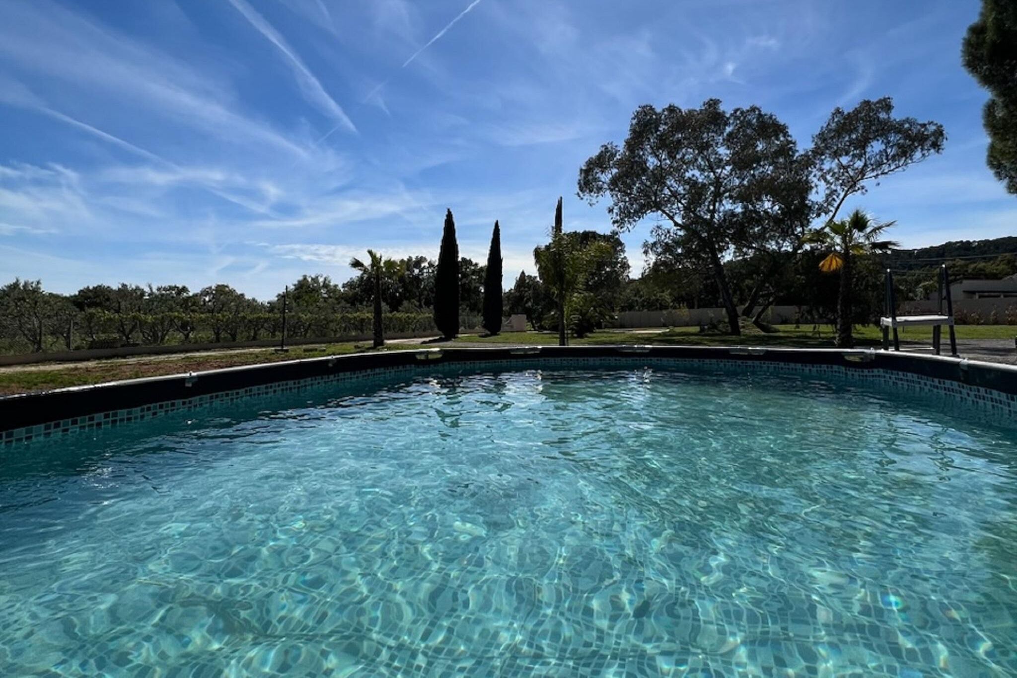 Una piscina azul cristalina con borde de azulejos, rodeada de árboles y un cielo despejado.