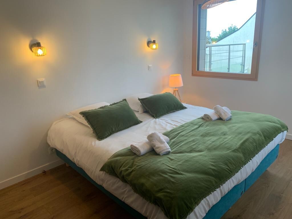 Una cama bien hecha con ropa de cama verde y blanca, dos lámparas fijadas a la pared y una ventana con vistas a un edificio.