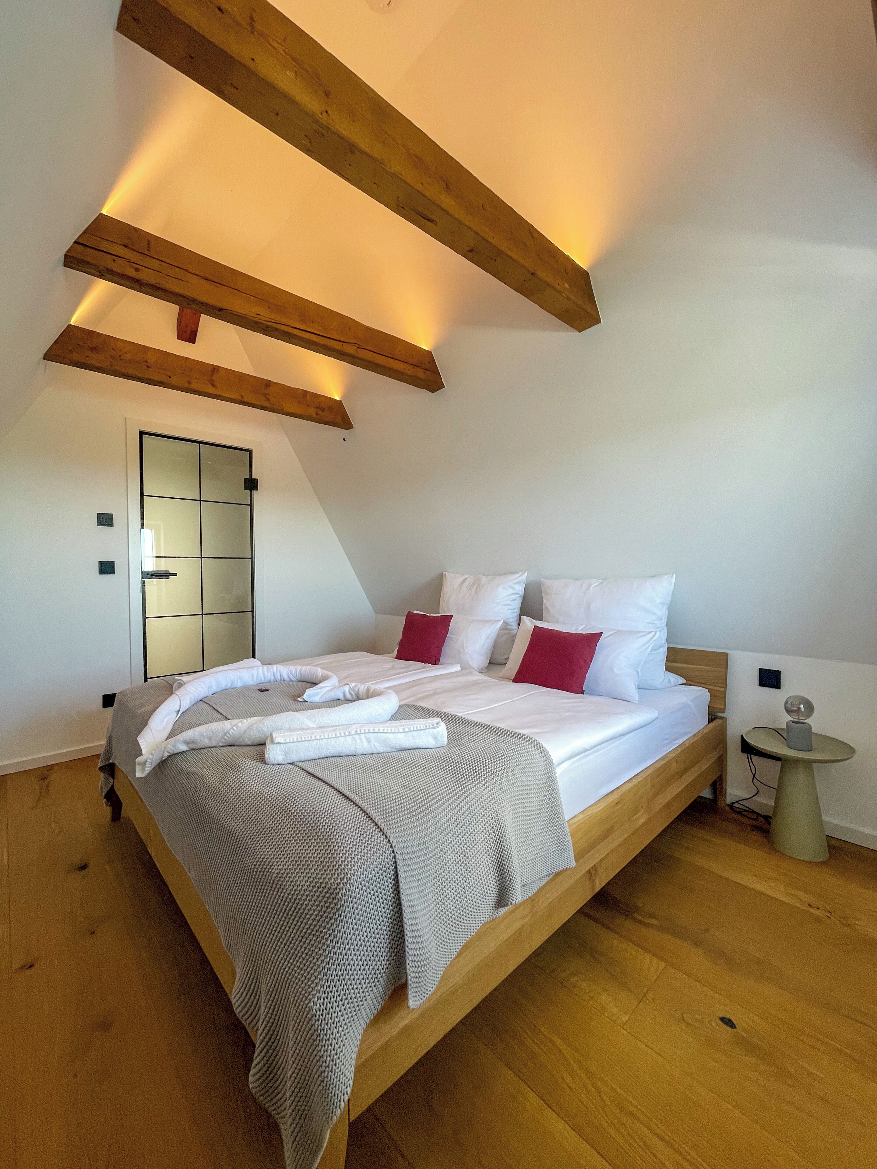 A bedroom with a wooden bed, white bedding, red pillows, a bedside table with a lamp, and a mirror on the wall.