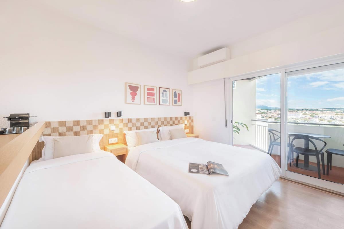 A hotel room with two beds, a checkered headboard, a balcony with a view, and a small table with chairs.