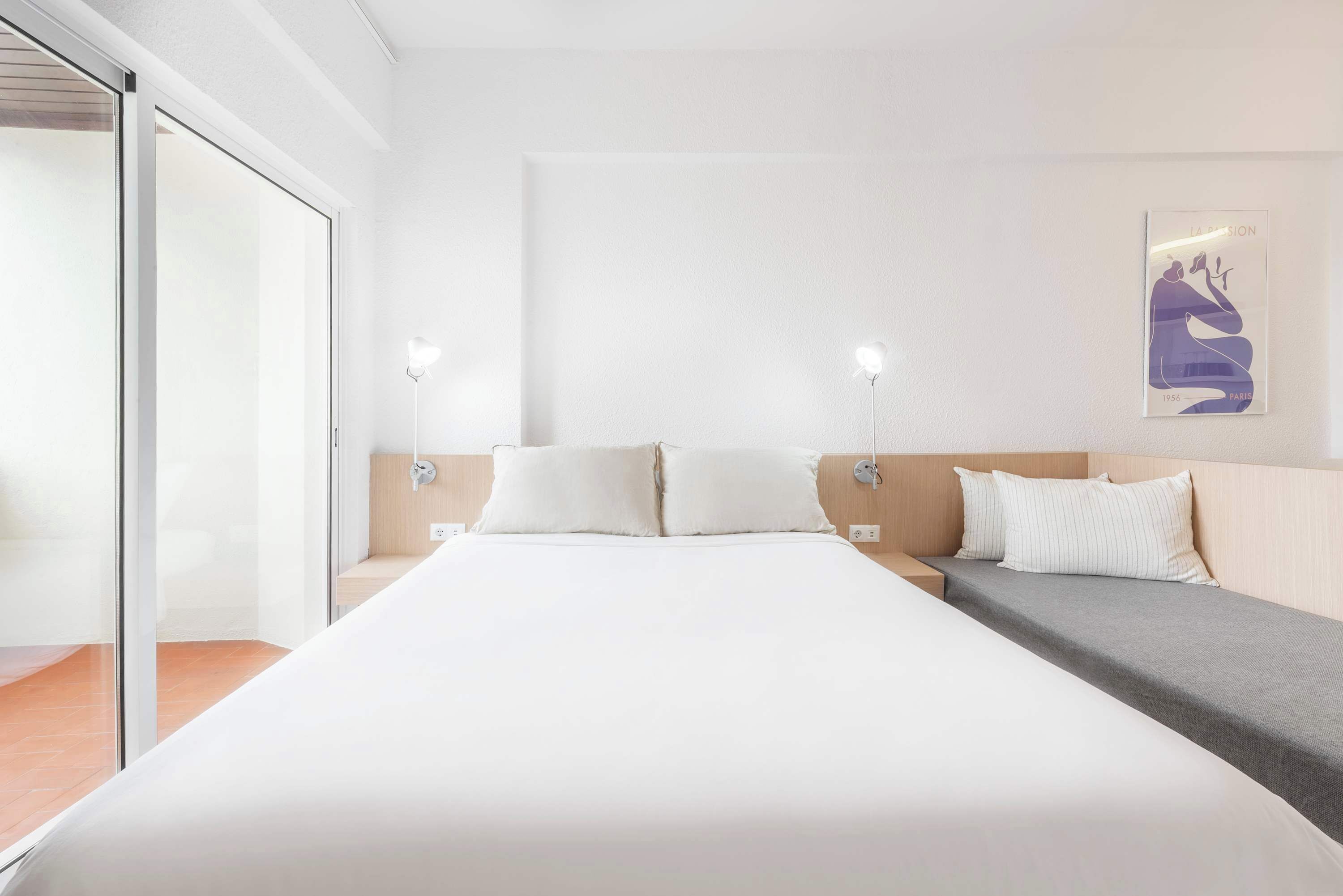 A modern hotel room with a large bed, a sofa, and a glass door leading to a balcony.