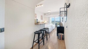 Un pasillo estrecho con pared blanca texturizada, suelo de madera y taburetes negros. Hay una pequeña zona de estar con un cojín colorido y un estante negro con objetos encima.