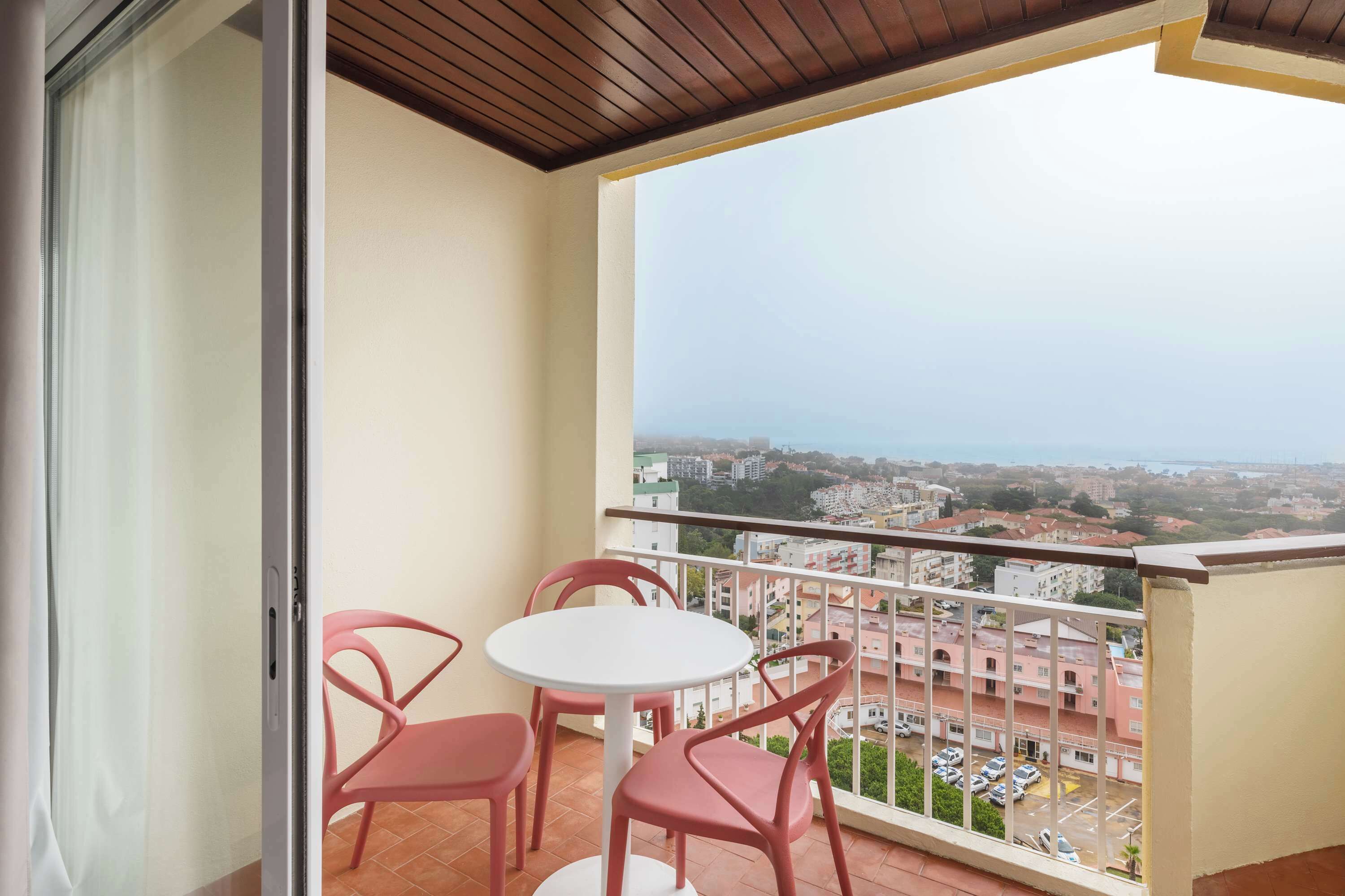 Un balcon avec une table blanche et quatre chaises roses, donnant sur un paysage urbain composé d’immeubles et d’un parking.