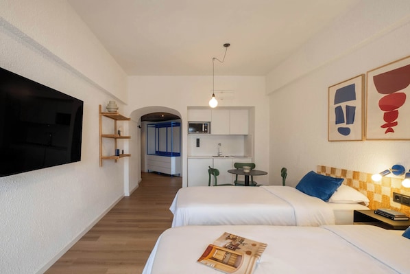 A hotel room with two beds, a TV, a kitchenette, and a hallway leading to another room.