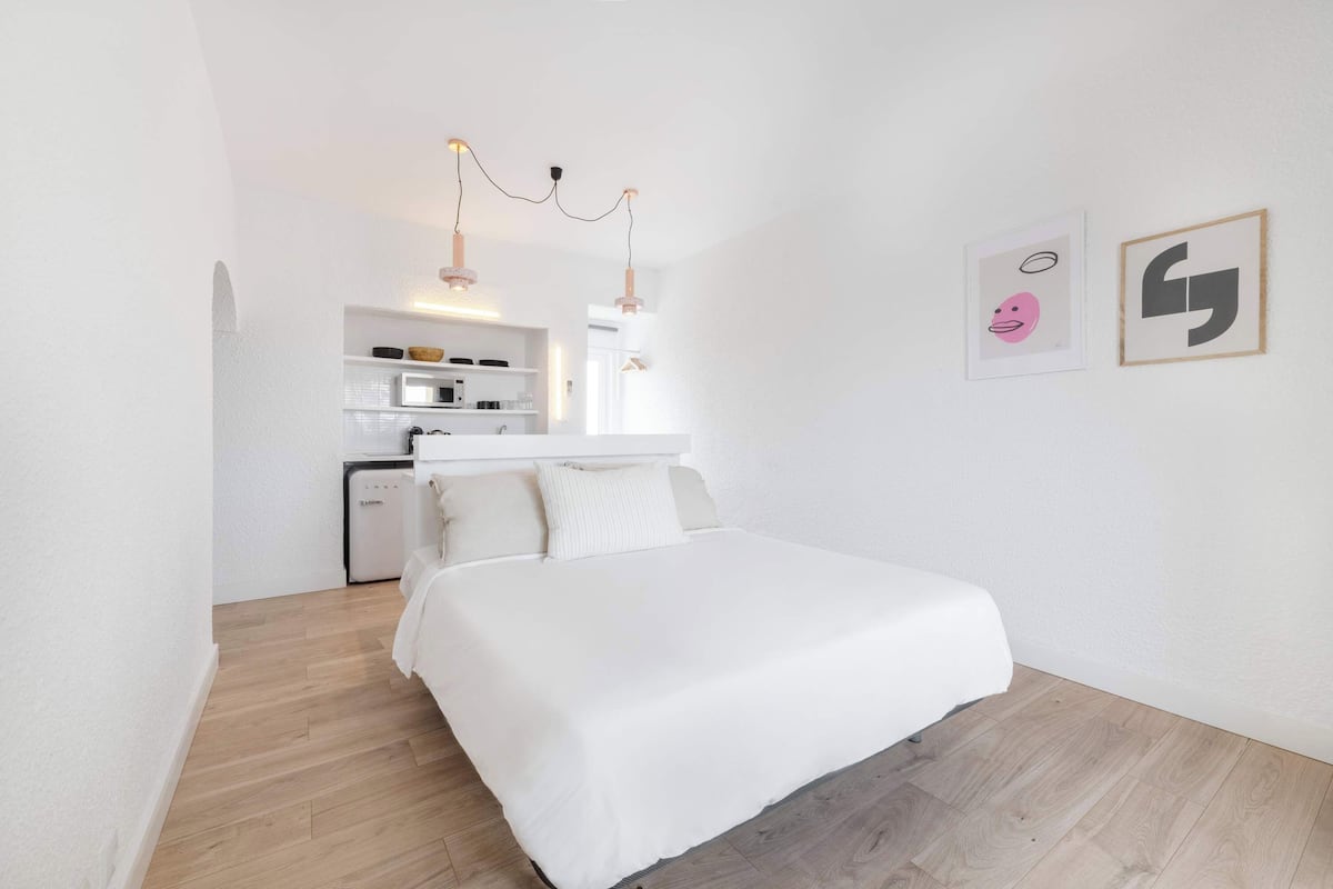 A modern, minimalist bedroom with a large bed, a small kitchenette, and a wooden floor.