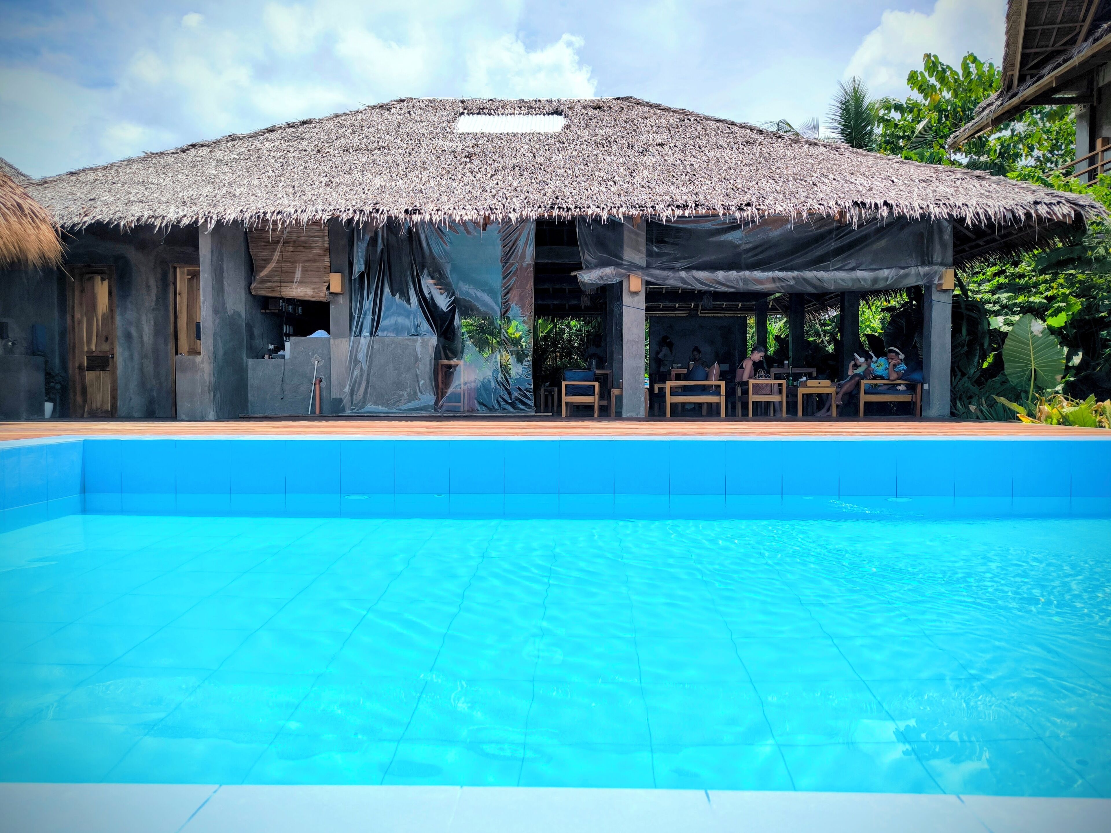 A poolside area with a thatched-roof structure, wooden chairs, and a clear blue pool.