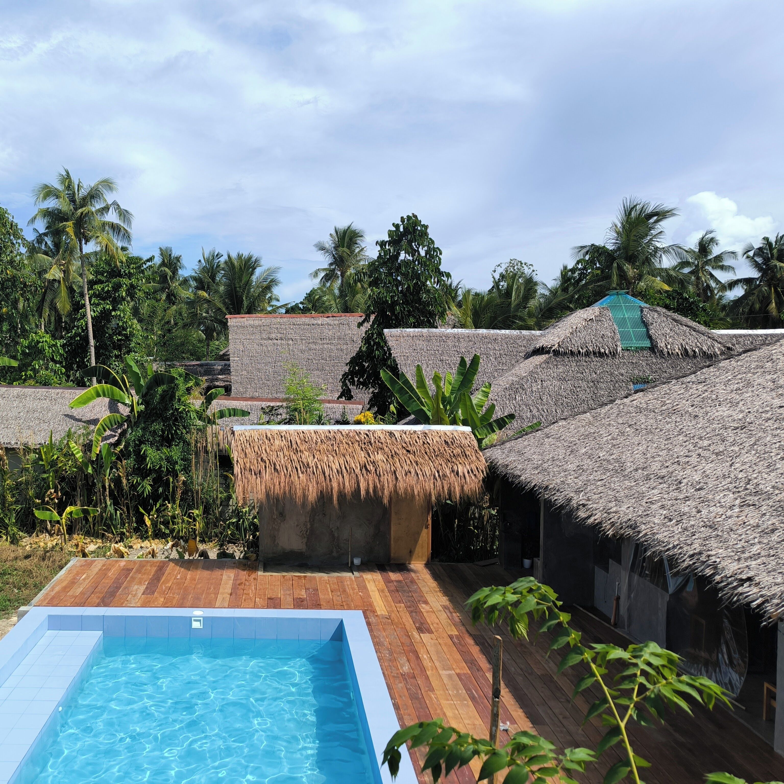 A pool area with a wooden deck, thatched-roof structures, and lush greenery.