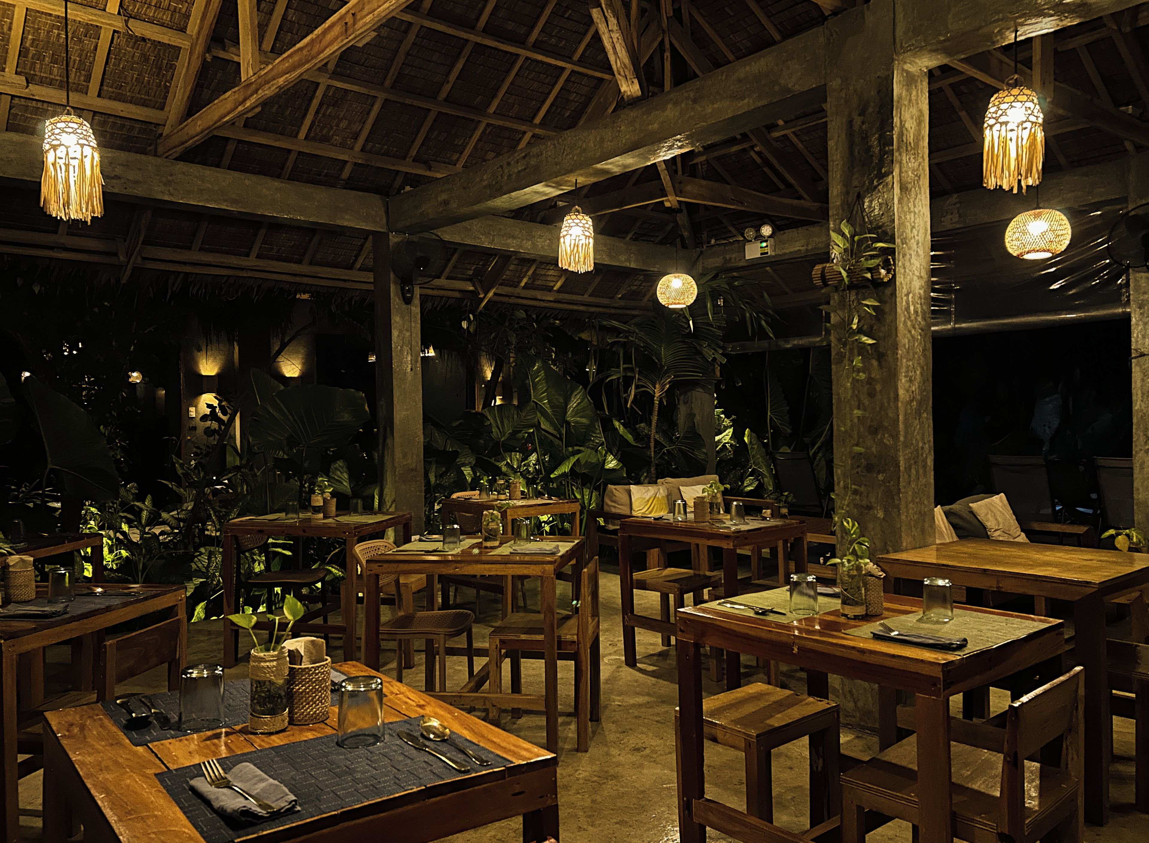 An indoor dining area with wooden tables and chairs, hanging pendant lights, and tropical plants.
