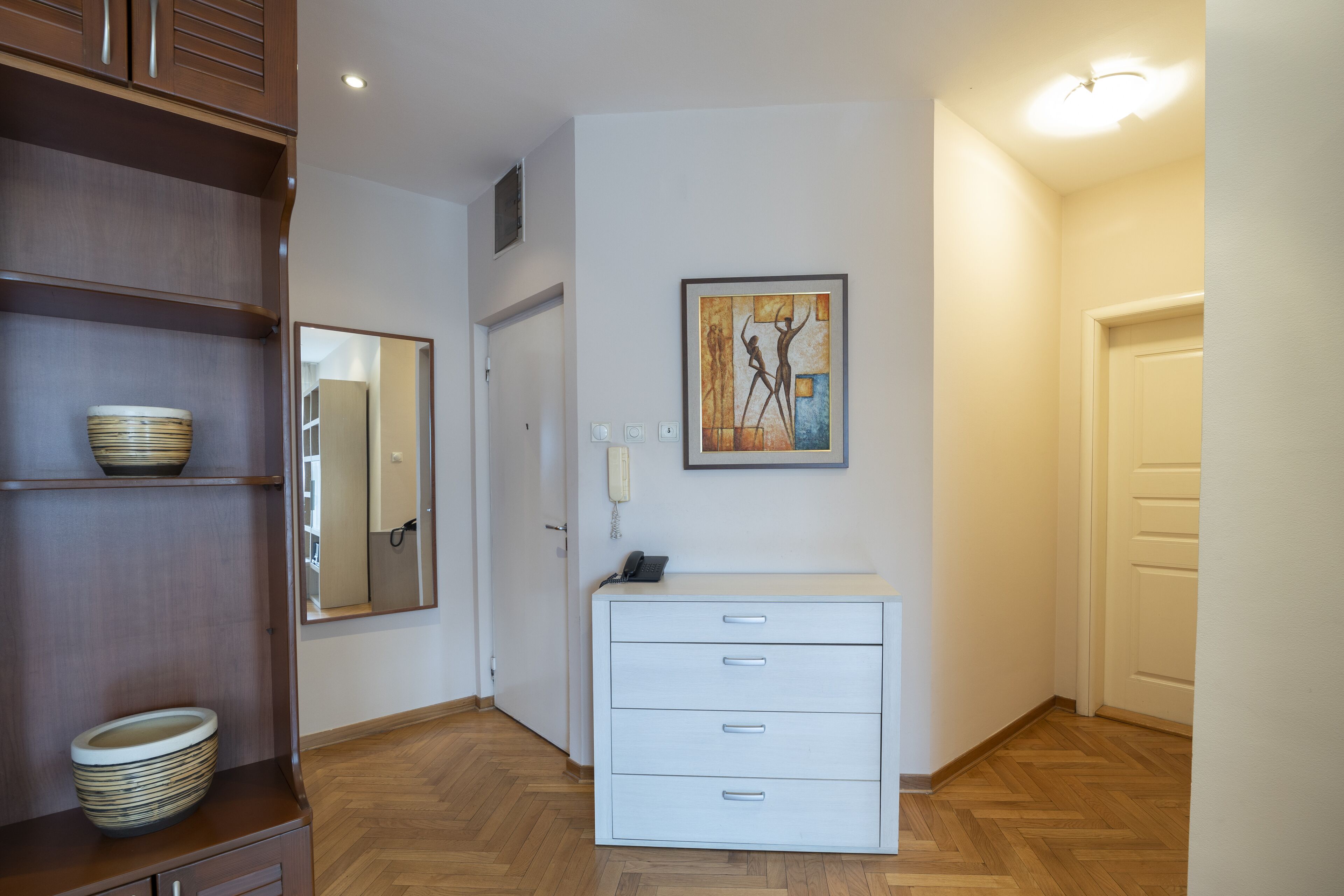 Une chambre d’hôtel avec un plancher en bois, une commode blanche avec des tiroirs, un miroir et un tableau accroché au mur.