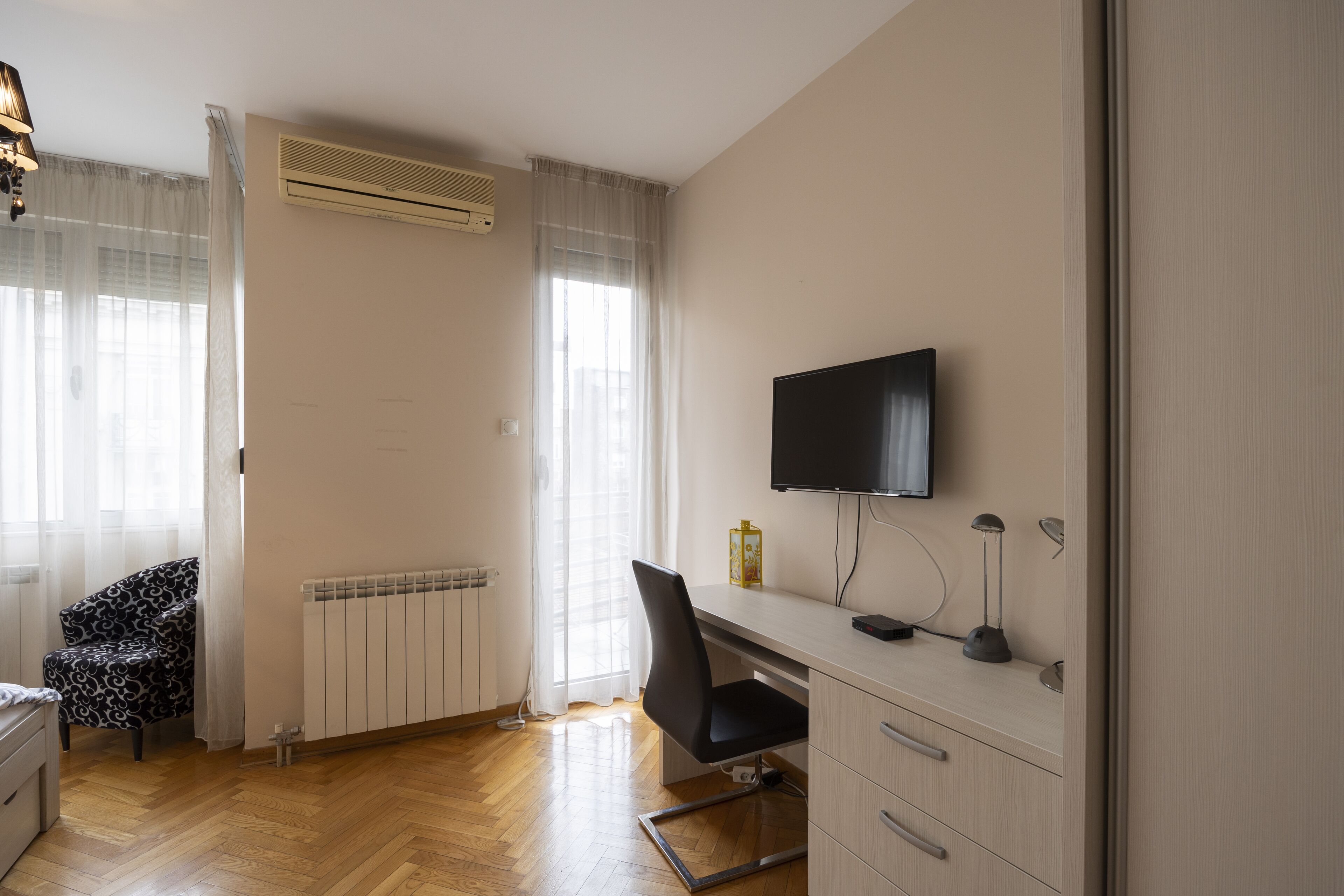 A room with a wooden floor, a desk with a chair, a TV mounted on the wall, and a radiator.