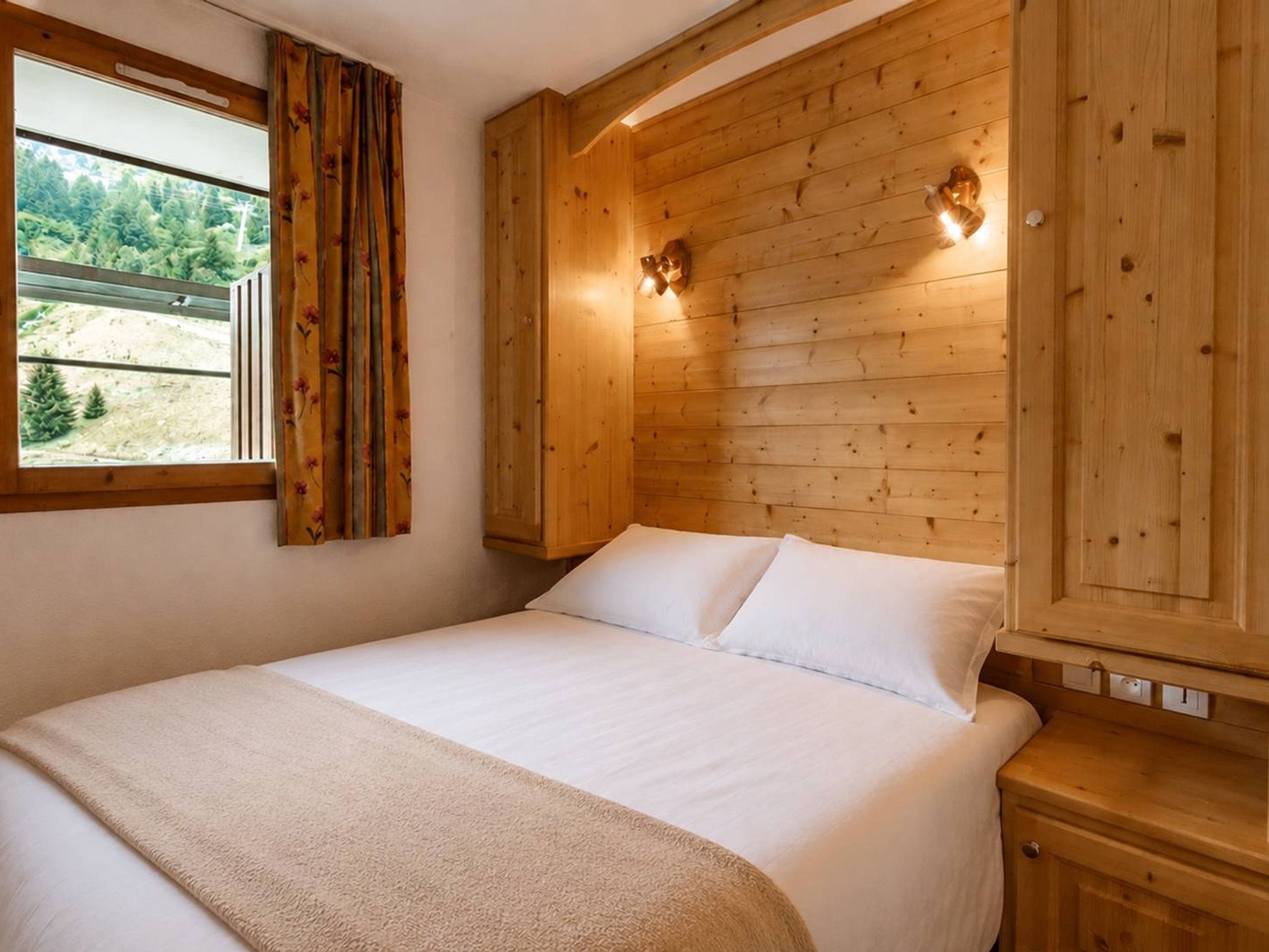 Une chambre aux murs lambrissés, avec un lit, une fenêtre avec des rideaux et des luminaires fixés au mur.