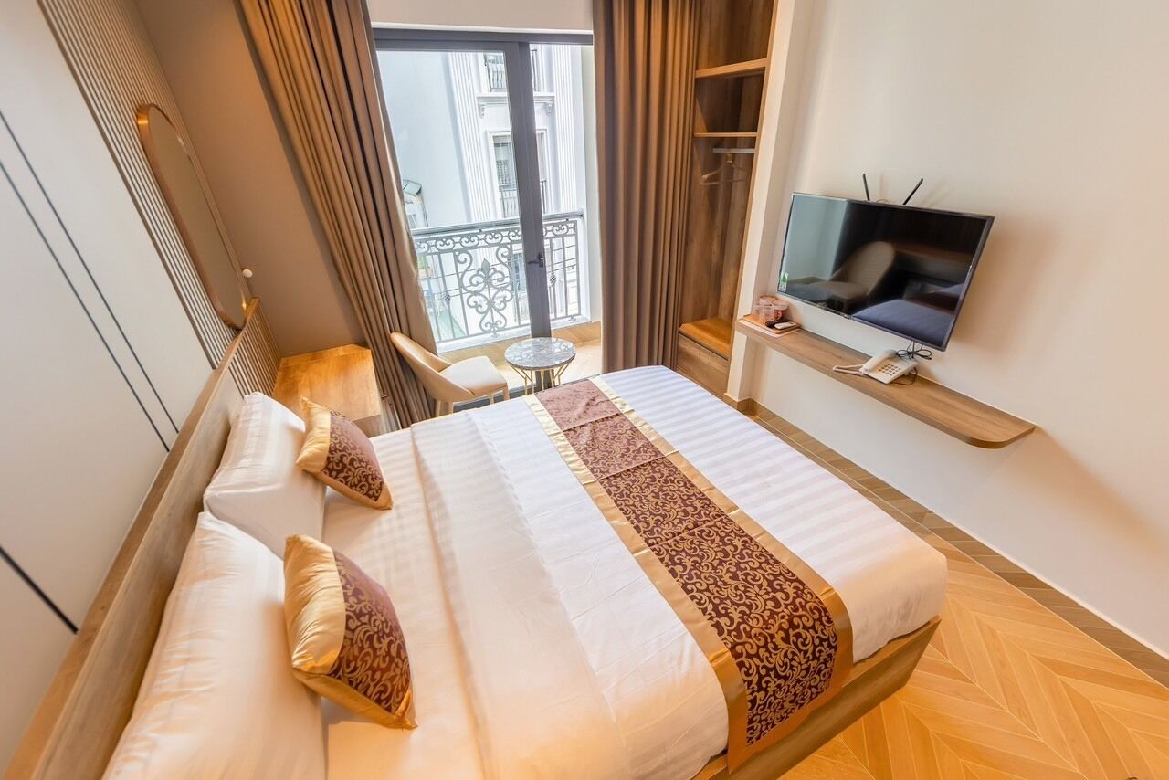 Habitació d'hotel amb llit, televisió, taula petita i balcó amb vistes a edificis.