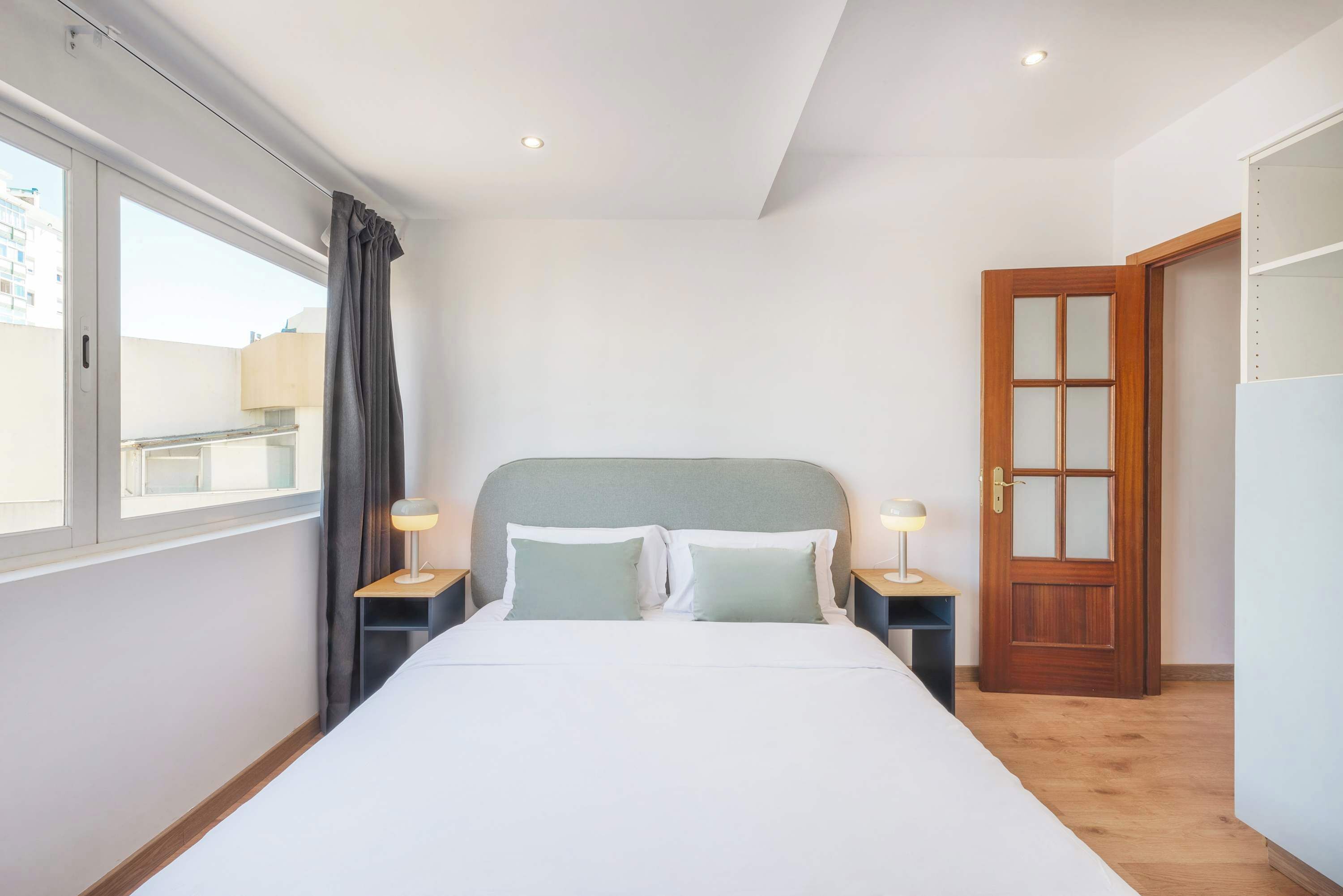 Une chambre à coucher avec un lit, deux tables de chevet, une fenêtre avec des rideaux, une porte en bois et une vue sur les bâtiments à l’extérieur.