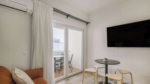 Een moderne woonkamer met een bank, een kleine ronde tafel met stoelen, een aan de muur bevestigde televisie en een schuifdeur naar een buitenruimte.