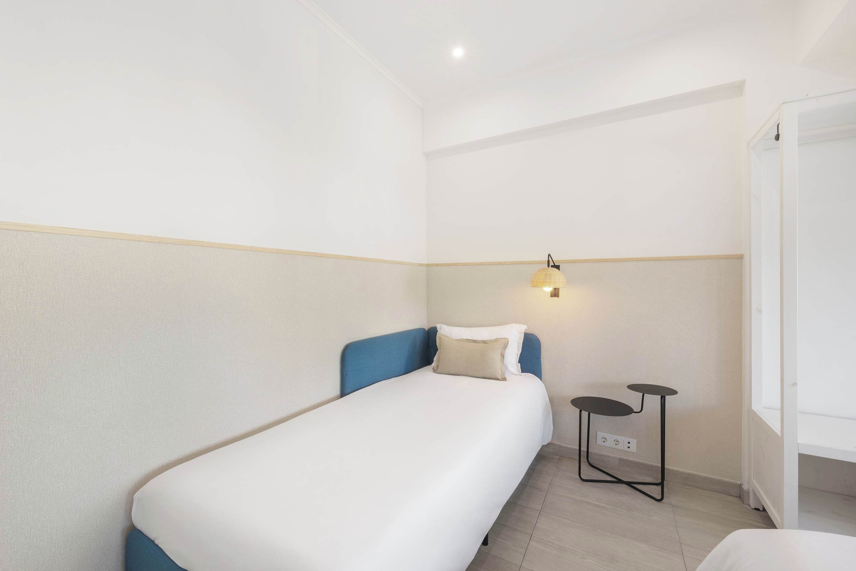 Un lit simple avec une tête de lit bleue et du linge de lit blanc, dans une petite chambre propre, équipée d’un interrupteur et d’une petite table.