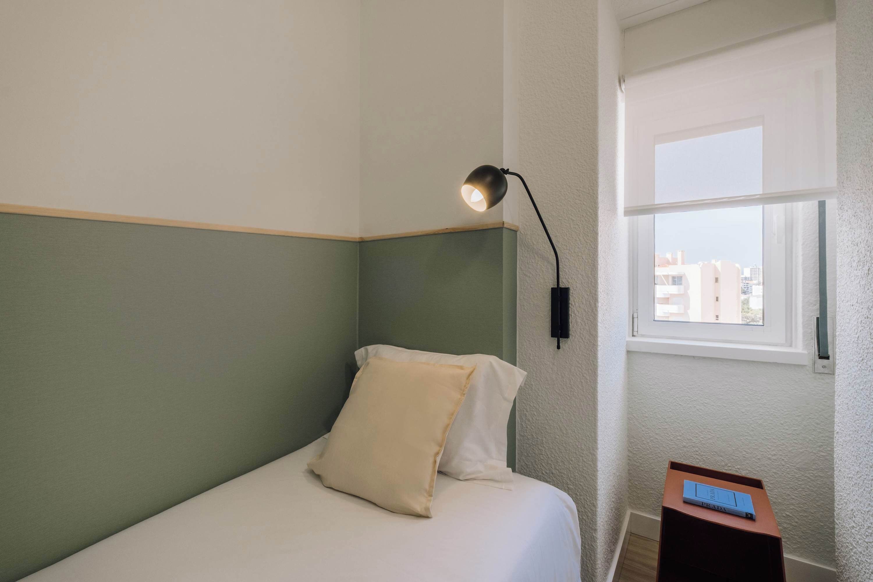 Una camera da letto piccola e ordinata con un letto singolo, un comodino con una lampada e una finestra vista edifici.