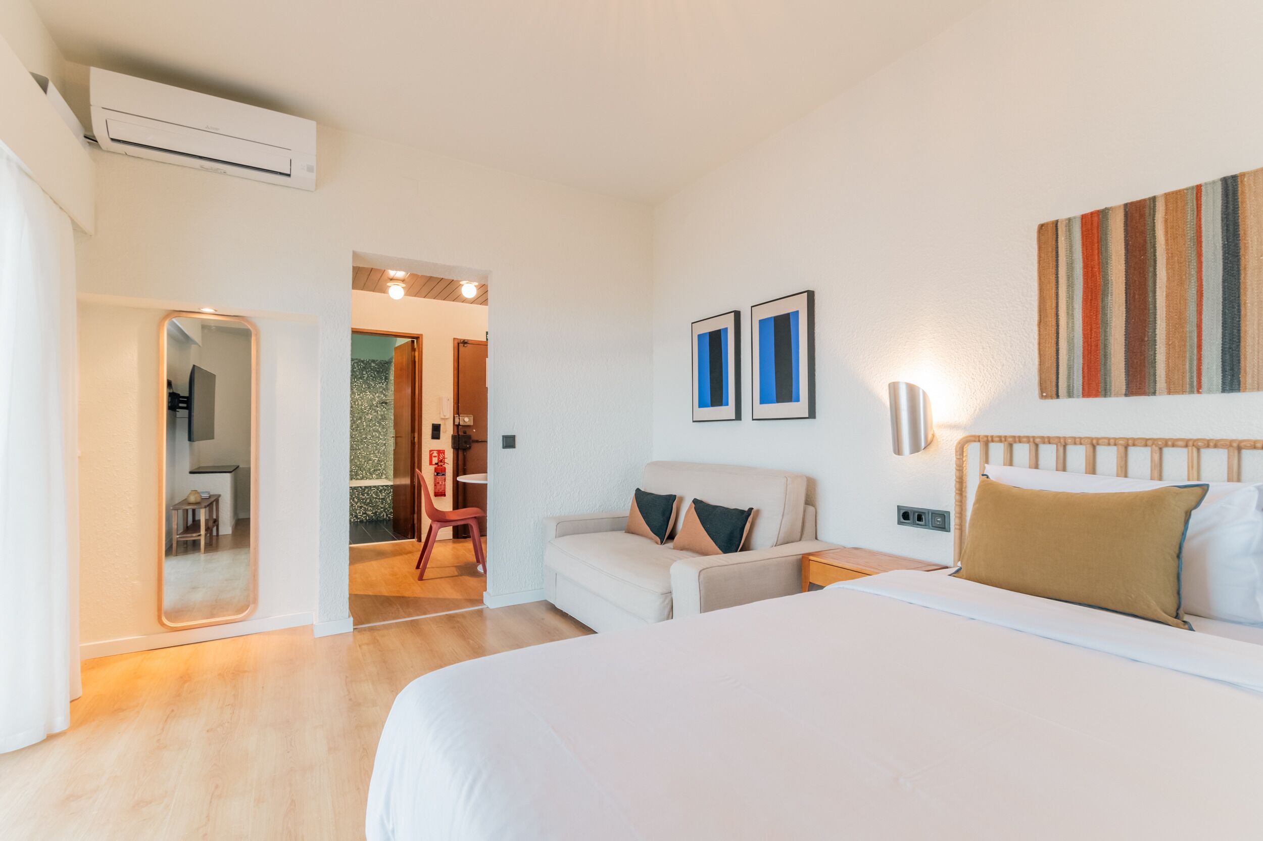 A hotel room with a bed, a sofa, a mirror, and a wall-mounted air conditioner.