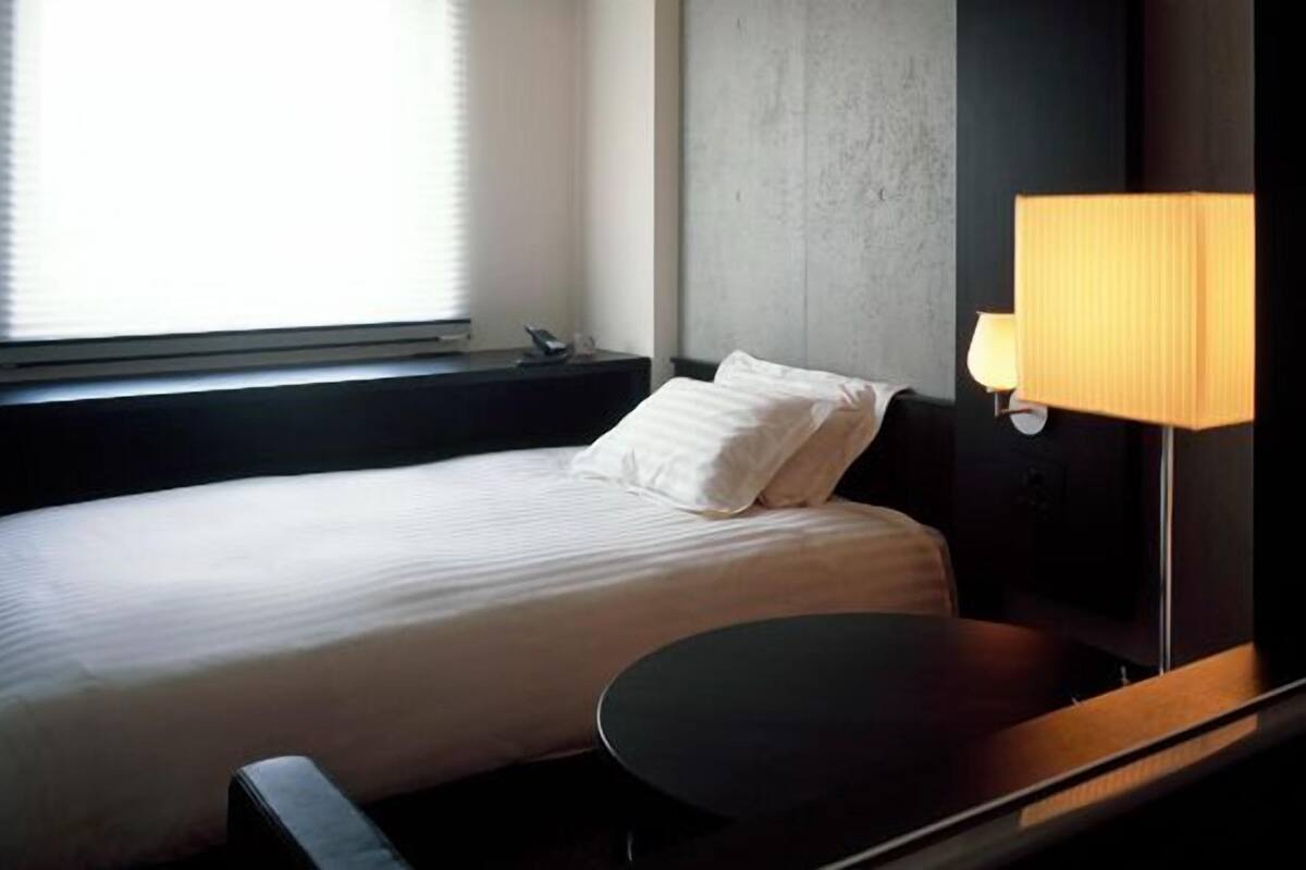 Una habitación de hotel con cama, mesita de noche, lámpara y ventana con persianas.