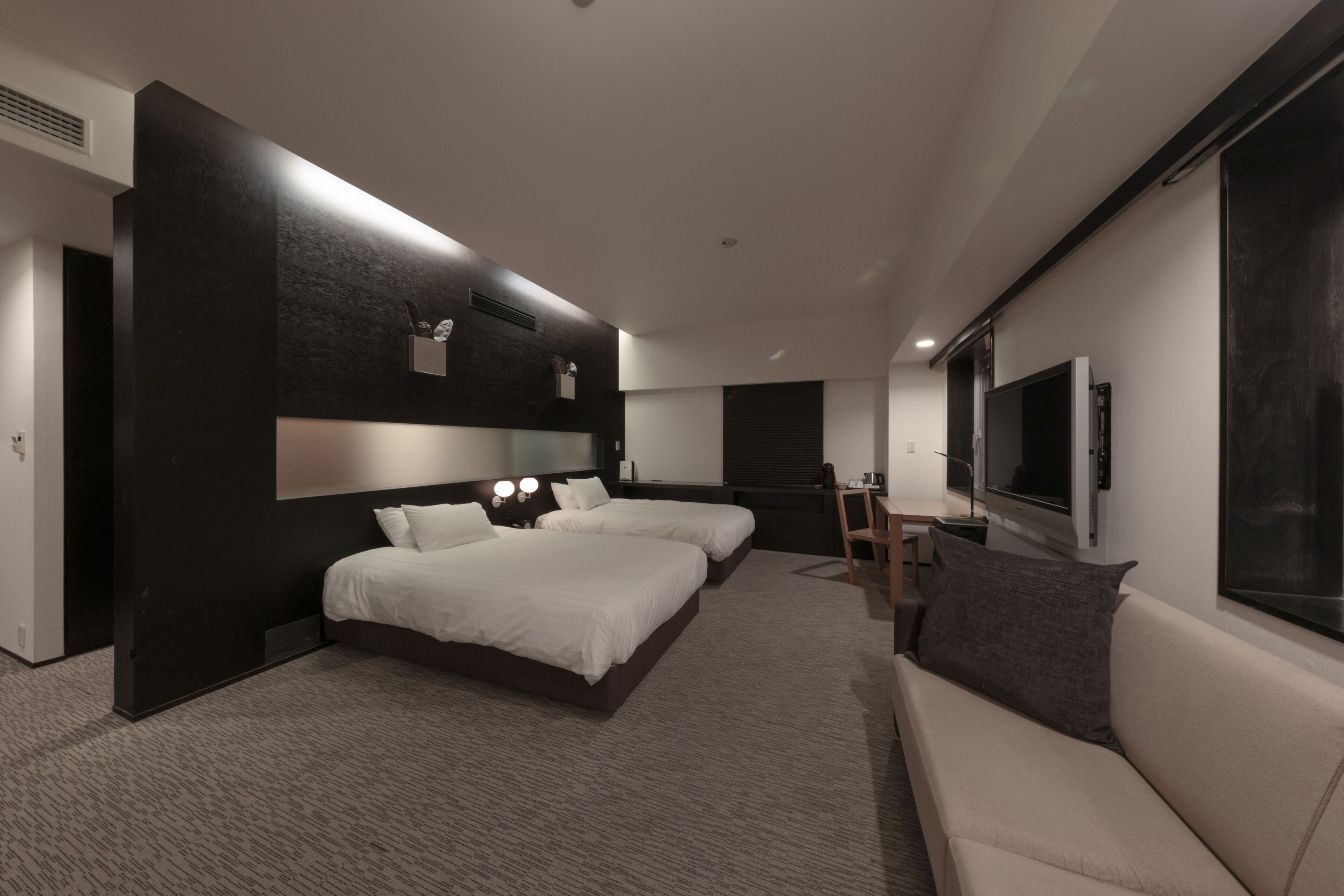 A modern hotel room with two beds, a sofa, a TV, and a desk.