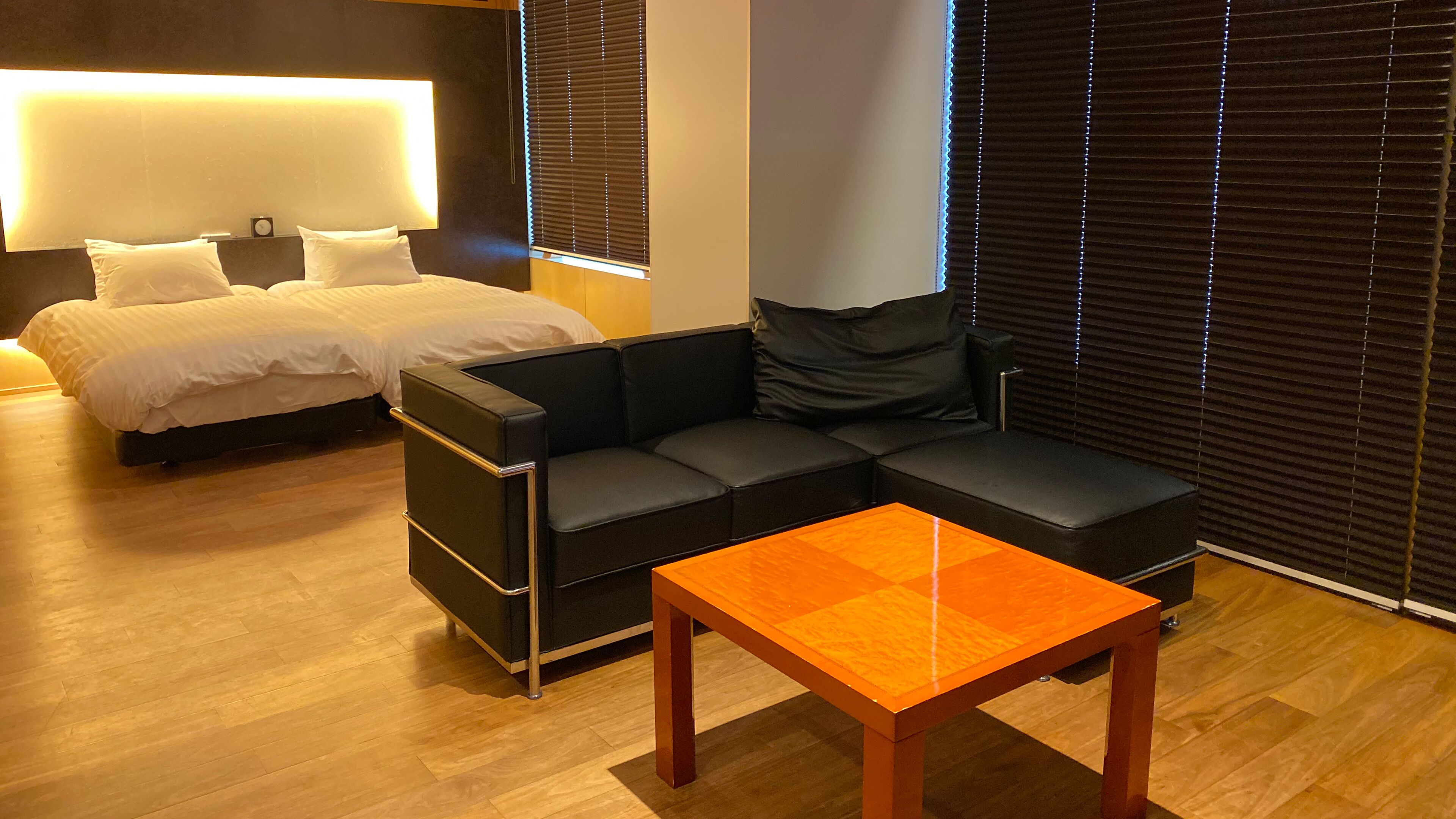 A modern hotel room with a bed, a black leather sofa, a wooden coffee table, and a large window with blinds.