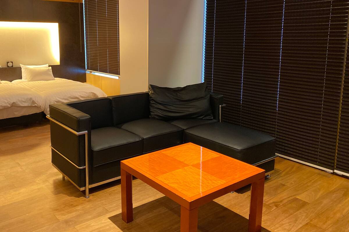 Una habitación de hotel moderna con una cama, un sofá de cuero negro, una mesa de centro de madera y un ventanal con persianas.