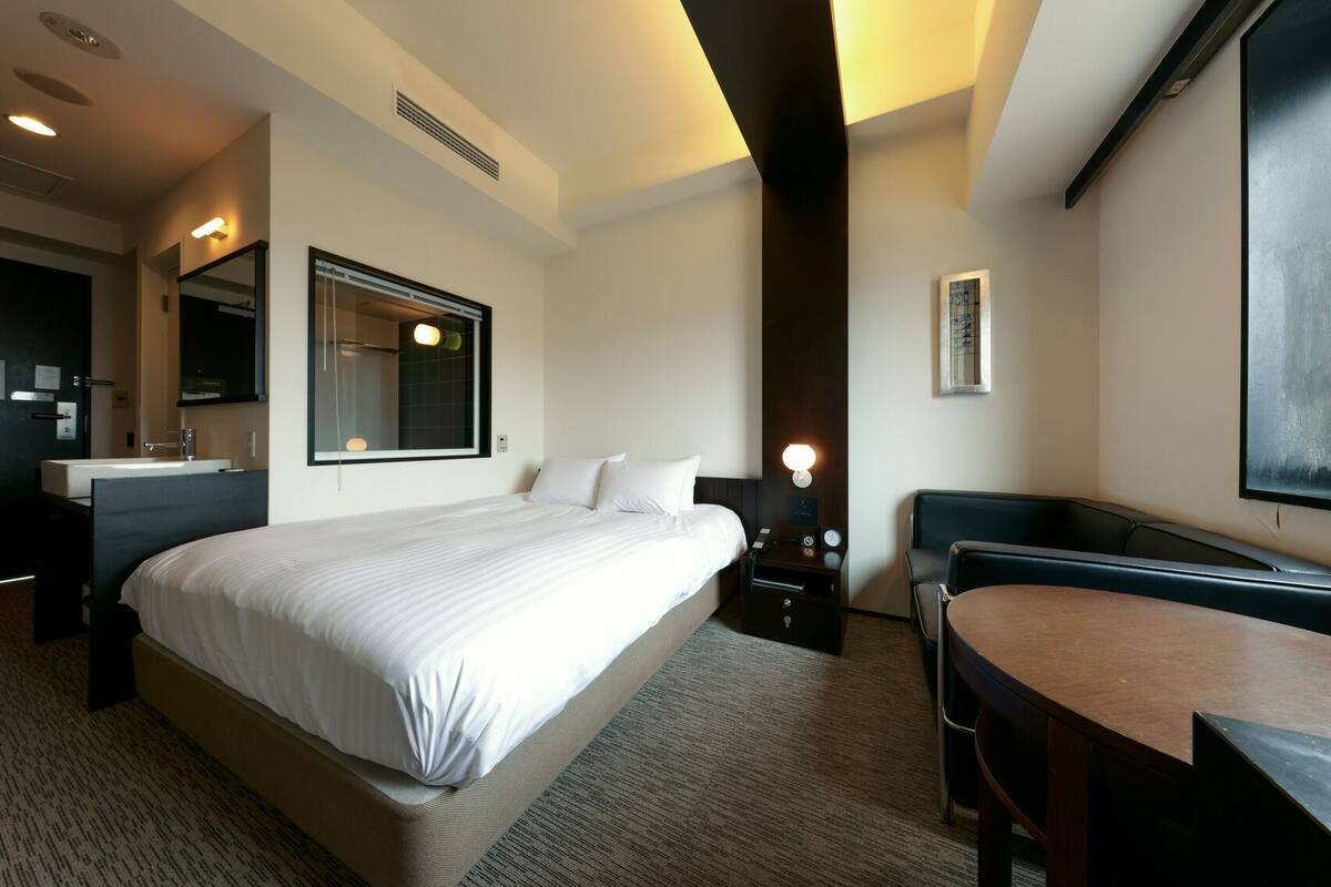 Habitación de hotel con una cama grande, un escritorio, una silla, una mesita de noche con lámpara, un espejo y una ventana.
