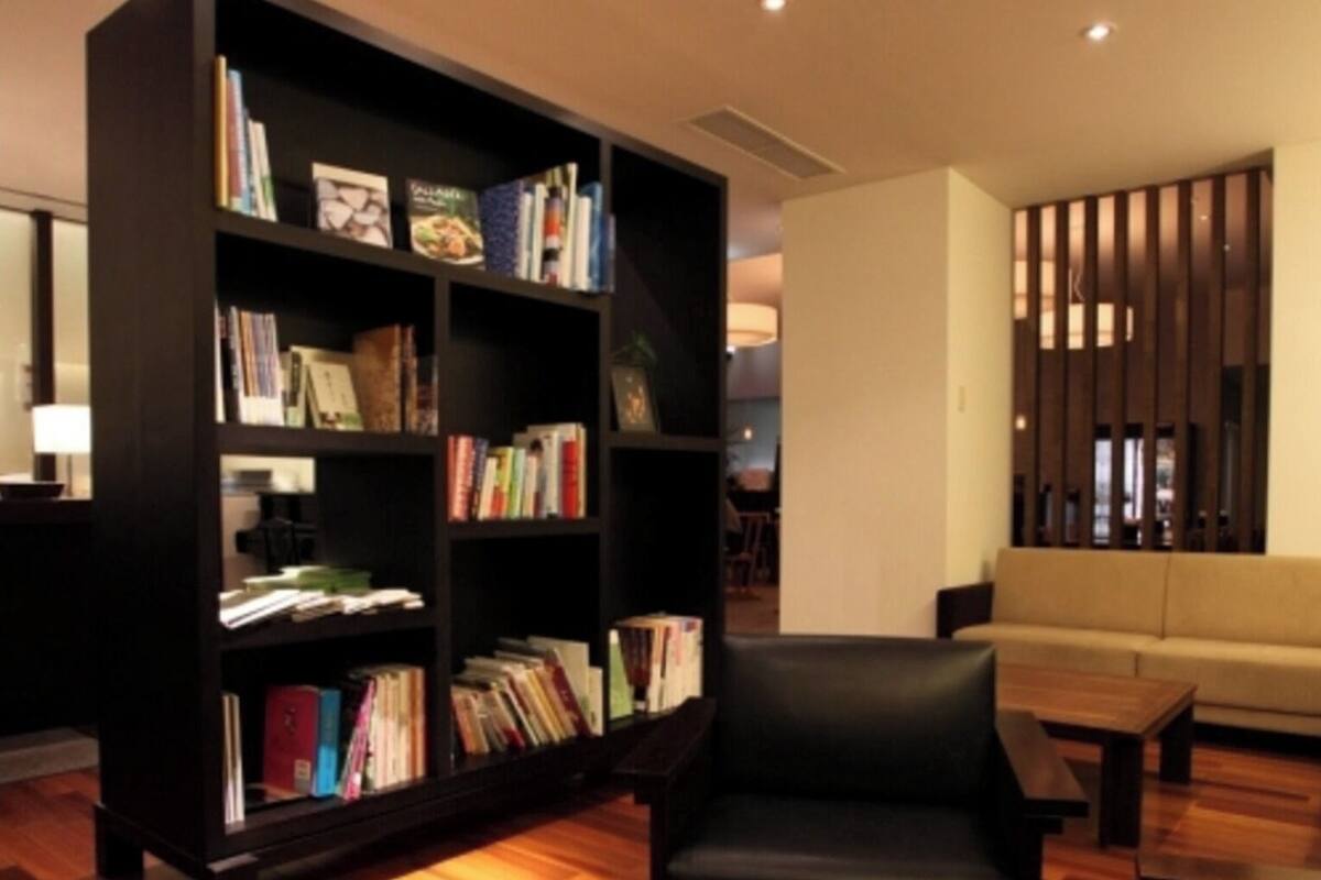 Una sala moderna con una amplia estantería negra llena de libros, un sillón de cuero negro, una mesa de centro redonda de madera y un sofá beige.