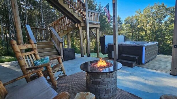 A wooden deck with a stone fire pit, a hot tub, and a wooden chair with cushions. - Hot Tub, Lake Front, NEW Game Rm, Fire Pit, Kayaks (Eureka Springs)