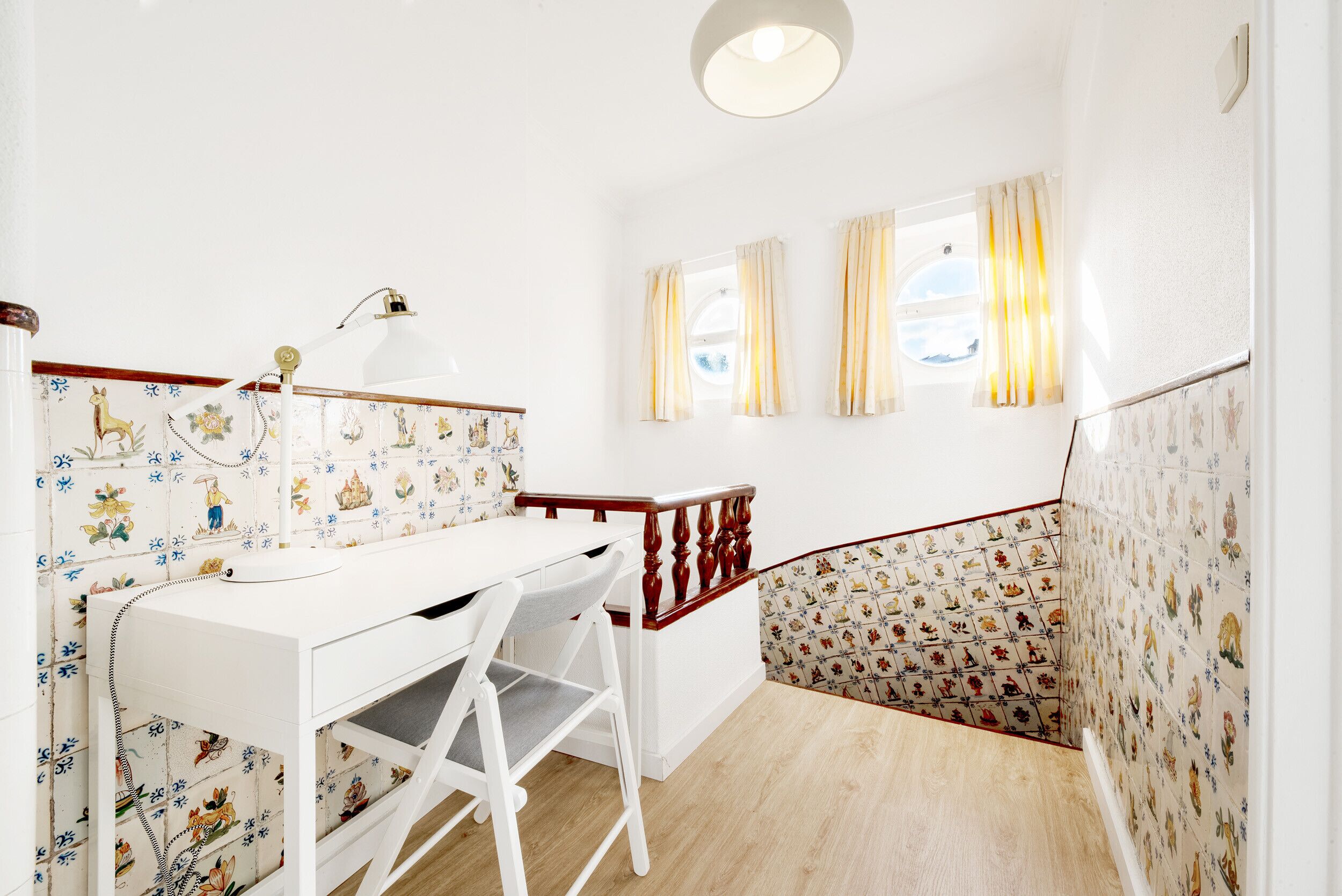 Una habitación con un escritorio blanco, sillas y una lámpara. Hay ventanas con cortinas y una escalera con papel tapiz estampado.