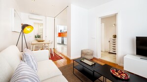 A modern living room with a sofa, coffee table, and a television. There is a dining area with a table and chairs, and a kitchen in the background. - Navegantes Townhouse by Olala Homes. (Cascais)
