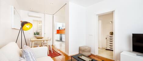 Una sala de estar moderna con un sofá, una mesa de centro y un televisor. Se observa un comedor con una mesa y sillas, y una cocina al fondo.