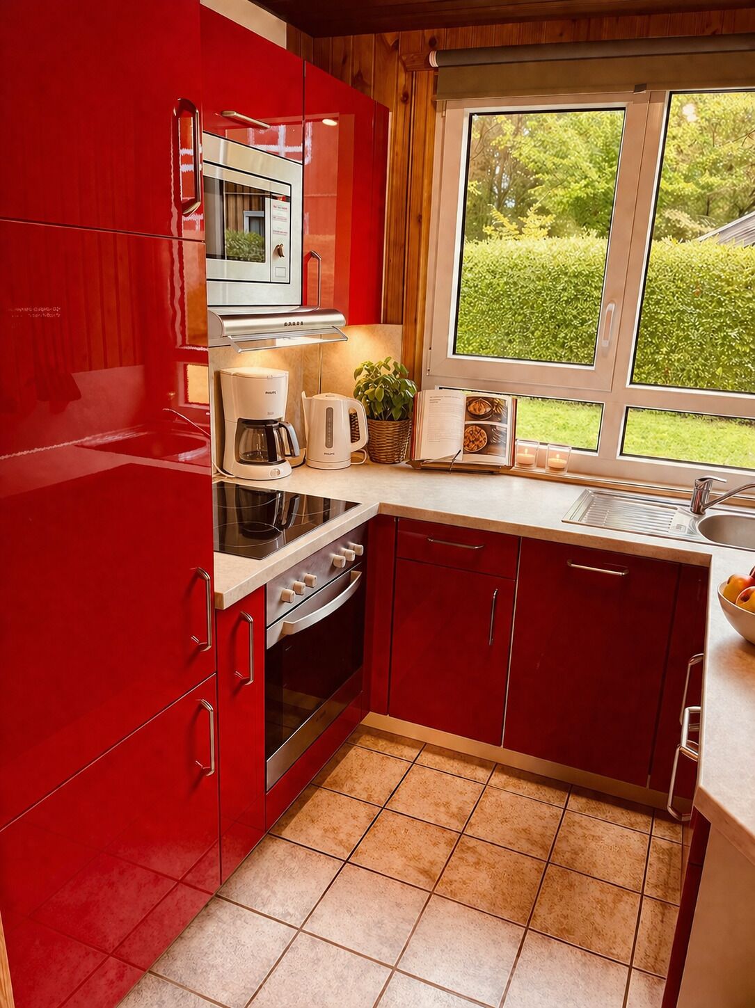 A kitchen with red cabinets, a stove, a coffee maker, and a window with a view of greenery.