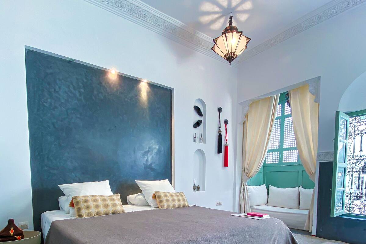 A bedroom with a large bed, a nightstand, a wall-mounted lamp, and a window with curtains.