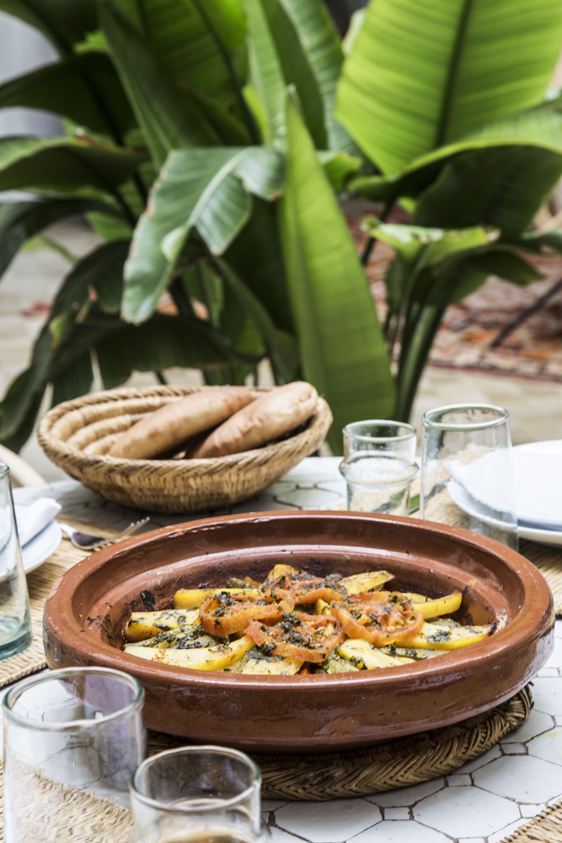 Un plat en terre cuite garni de légumes coupés en tranches, un panier de pain et un tapis tissé posé sur une surface carrelée.