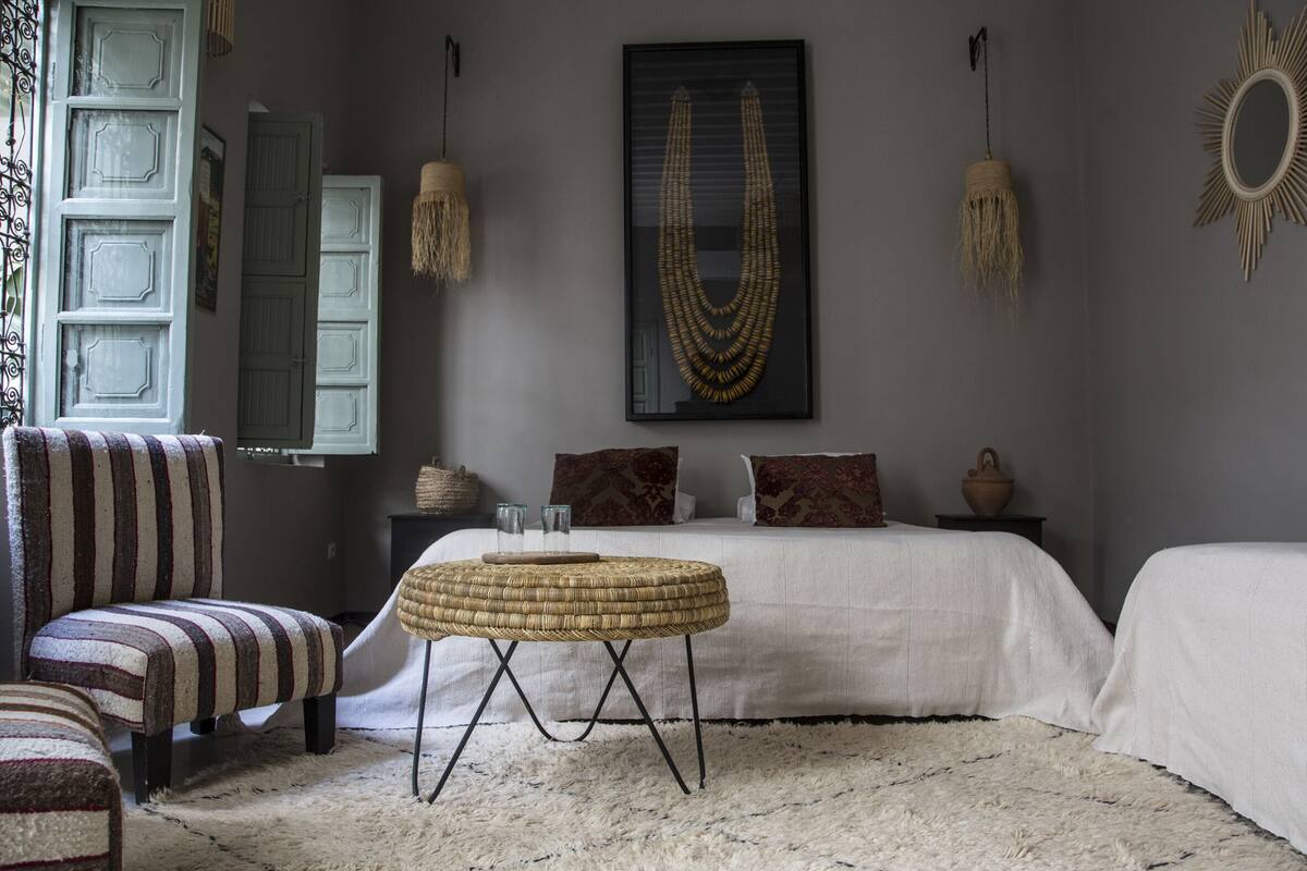A bedroom with a striped armchair, a round woven table, a bed with white bedding, and a wall-mounted mirror.