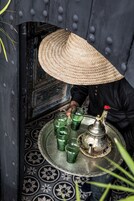 Una persona sirve bebidas en una bandeja con una jarra metálica tradicional y vasos, en un ambiente con baldosas de suelo estampadas y una puerta decorativa.