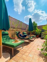 Un espacio lounge al aire libre bañado por el sol, con sillas acolchadas verdes y sombrillas, rodeado de macetas con plantas y una valla de celosía de madera.