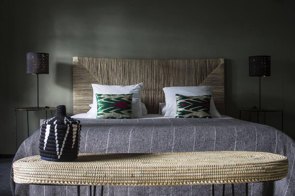 A neatly arranged bedroom with a large bed, two bedside lamps, a woven pouf, and a textured wall headboard.