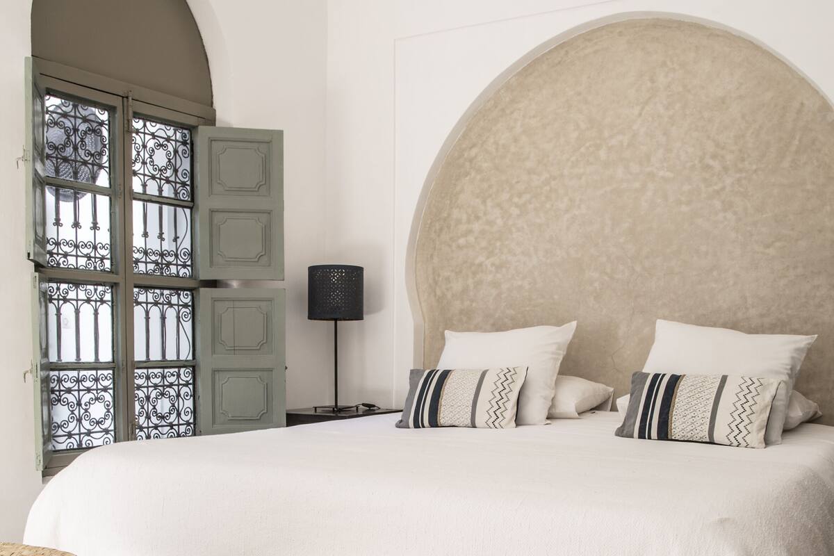 A bedroom with a large bed, a round headboard, a patterned rug, a woven chair, and a window with metal bars.