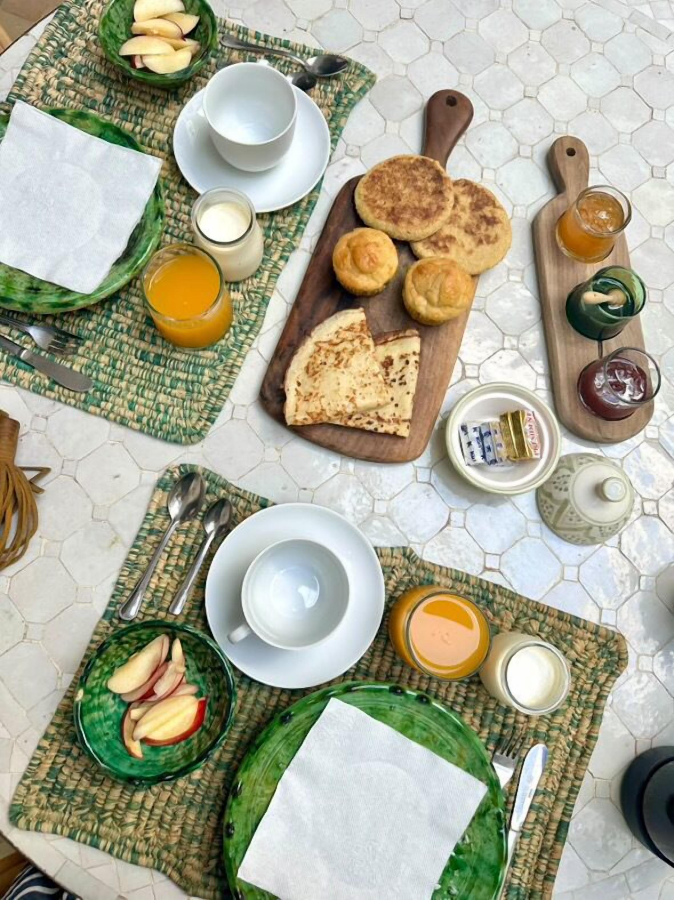 A breakfast setting with a variety of food items, including pancakes, apple slices, and beverages, arranged on a table with a hexagonal tile floor.