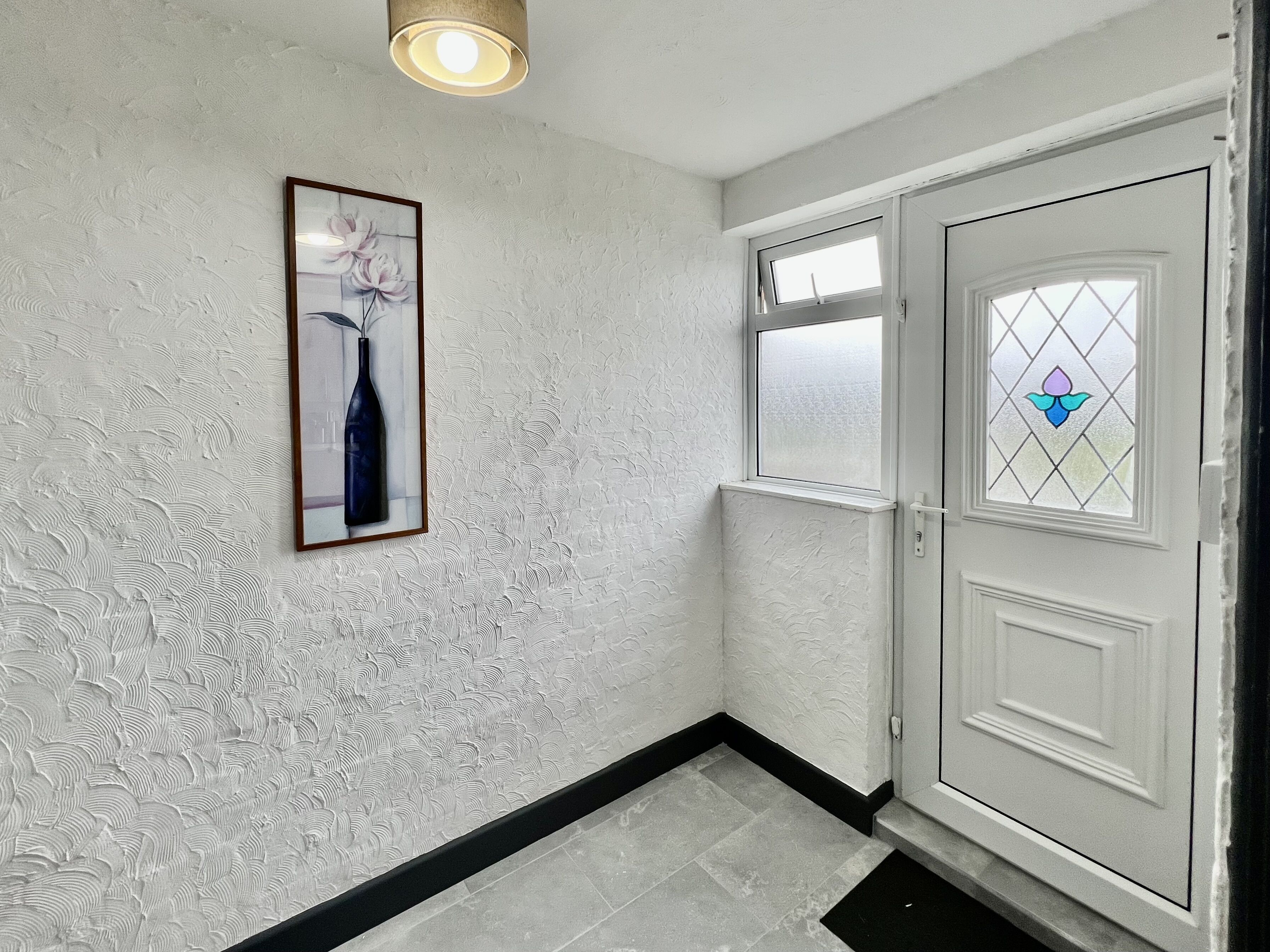 A hallway with textured walls, a framed picture of a vase, a door with a stained glass window, and a light fixture.