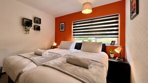 A bedroom with a bed, a bedside table with a lamp, a window with blinds, and a wall-mounted picture frame. - Havenwood House (Nottingham)