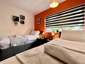 A hotel room with two beds, a window with blinds, a bedside table with a lamp, and a small shelf with plants. - Havenwood House (Nottingham)