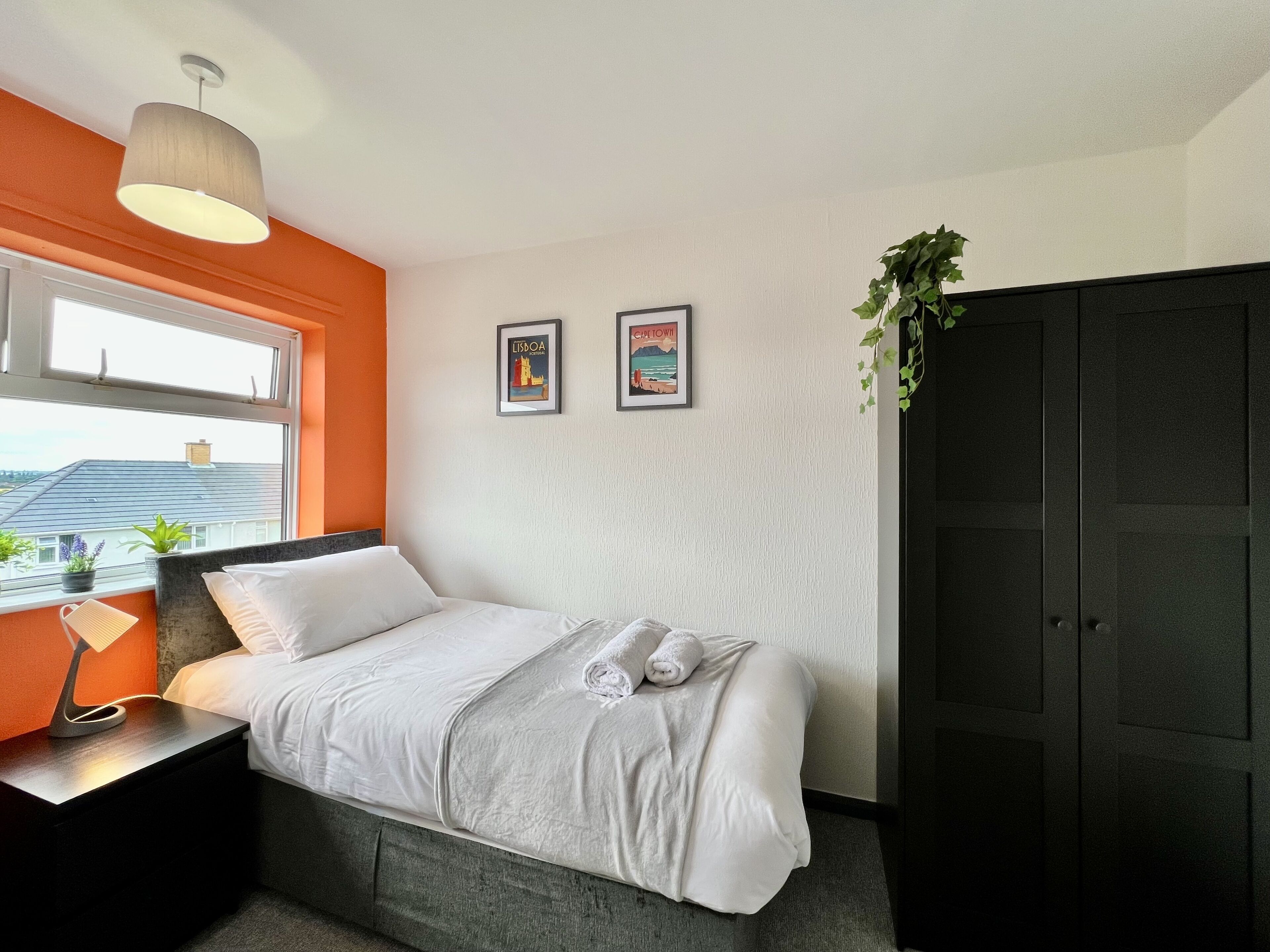 Een hotelkamer met een bed, nachtkastje, lamp, raam met uitzicht, twee ingelijste foto's aan de muur en een donkere garderobe.